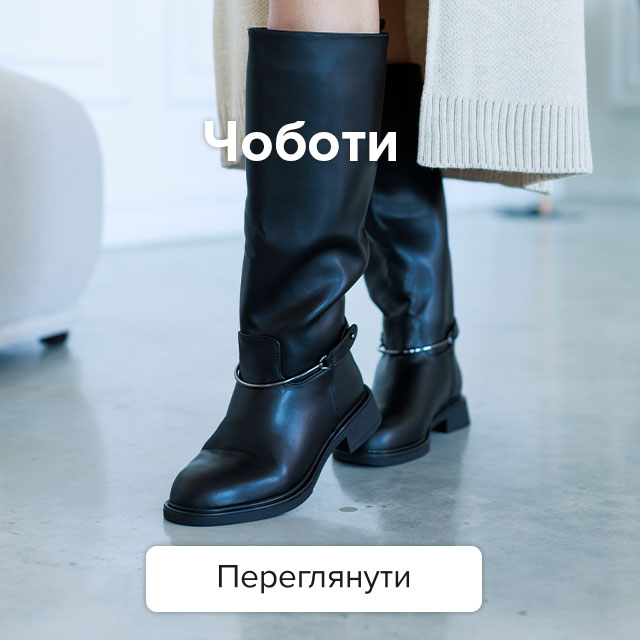 Високі жіночі чорні шкіряні чоботи на плоскому ходу з декоративним металевим ремінцем на щиколотці в поєднанні зі світлим довгим кардиганом.