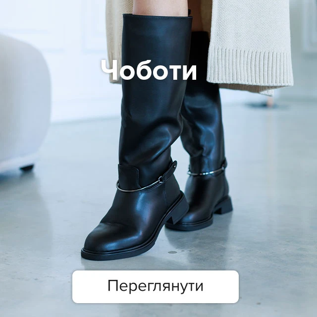 Високі жіночі чорні шкіряні чоботи на плоскому ходу з декоративним металевим ремінцем на щиколотці в поєднанні зі світлим довгим кардиганом.