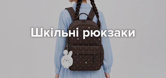 Категорійний банер «Шкільні рюкзаки»: дівчина у сірому с світшоті та джинсах тримає бежево-коричневий шкільний рюкзак із брелоком-іграшкою