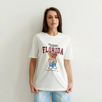 Дівчина у білій вільній футболці з принтом ведмедя та написом «FLORIDA» та світло-блакитних джинсах стоїть на світлому тлі