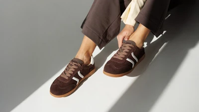 Женские коричневые замшевые кеды с белыми полосками и светлой резиновой подошвой, на ноге в луче света