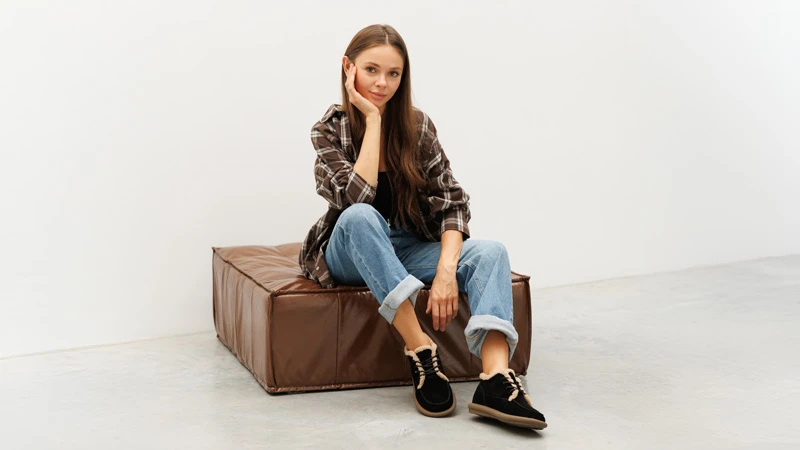 Жінка в чорних уггах у повсякденному образі: джинси з підворотами й картата сорочка, сидить на коричневому пуфі.