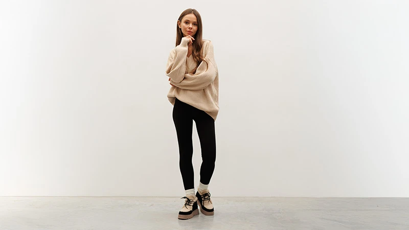 Жінка в жіночих уггах із бежевим хутром, чорні легінси та бежевий оверсайз светр, повний зріст на світлому фоні.